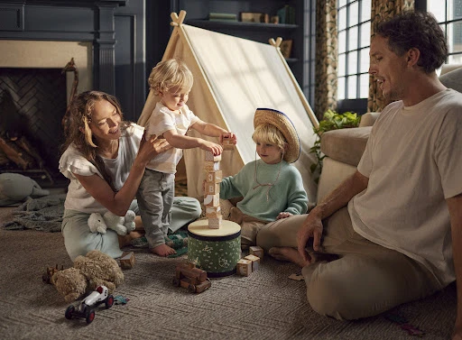 A family of three, a mother, father, and two children, play with wooden blocks inside a tent indoors, creating a heartwarming and playful scene.