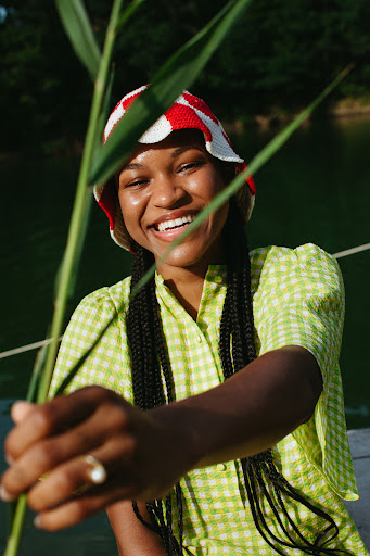 A woman smiles in a portrait outside, wearing a colorful hat and green dress, holding tall grass in front of a blurred lake background.