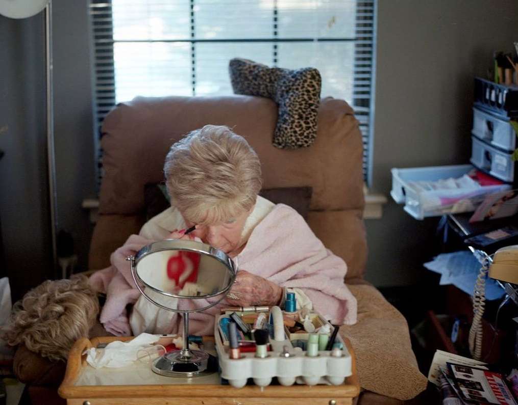 Elderly woman in a peach-colored robe applies makeup at a vanity table with a mirror, various cosmetics, and personal items in a home setting.