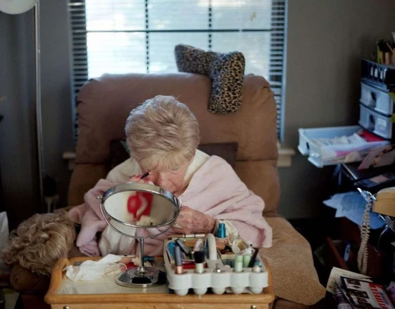 Elderly woman in a peach-colored robe applies makeup at a vanity table with a mirror, various cosmetics, and personal items in a home setting.
