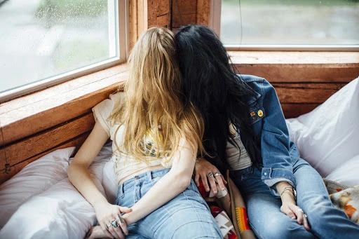 Two women with blonde and black hair sitting on a bed in front of a window in a rustic cabin with wooden walls, wearing denim jackets.