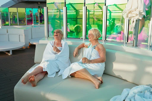 Two smiling elderly women sit wrapped in towels on a white couch. They're holding drinks and appear to be in a resort setting.