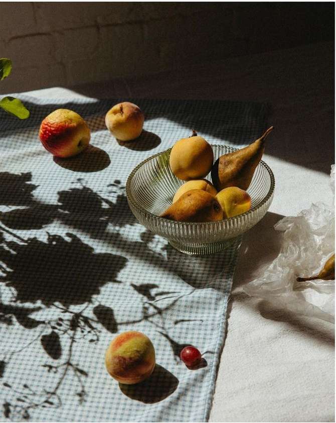 Still life featuring a glass bowl of pears, apples, and peaches on a blue gingham tablecloth with shadows of leaves and diffused lighting.
