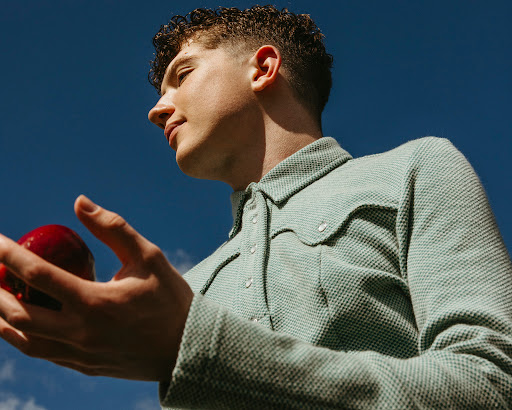 Low-angle view of a young adult male in a collared shirt holding a red apple against a bright blue sky with some clouds.