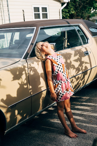 Child leaning against a vintage car on a sunny day, wearing a patterned dress and barefoot, with leaf shadows on the vehicle.