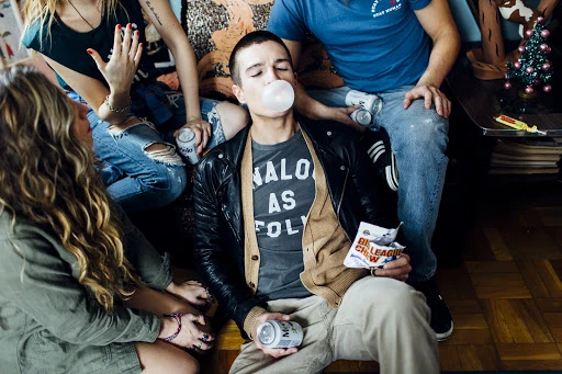 Group of young adults indoors, seated together with casual attire, drinking from cans and blowing bubbles, creating a relaxed, social atmosphere.