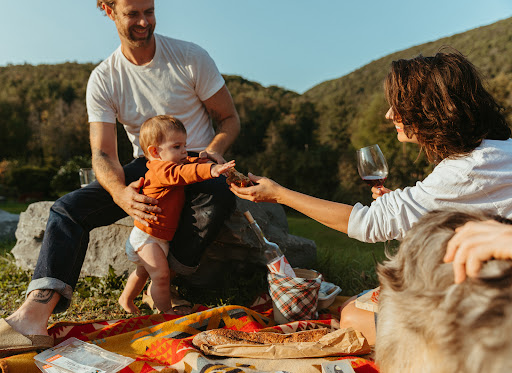 Family enjoying a picnic on a blanket outdoors with a baby, woman with wine, and rolling green hills in the background on a sunny day.