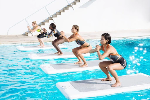A group of four women exercising in a pool on floating mats. They are doing squats and wearing swimsuits in an outdoor setting with white walls.