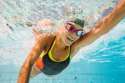 Underwater shot of a smiling adult woman swimming in a pool wearing a swim cap and goggles, showcasing fitness and an active lifestyle.