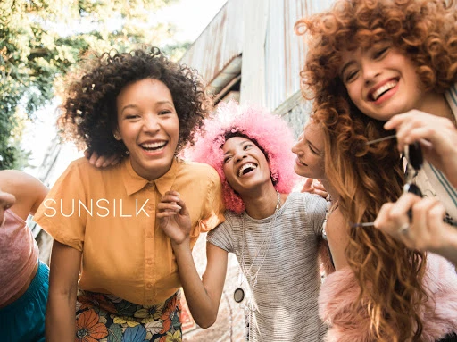 A group of diverse young women are laughing and smiling together outside while promoting Sunsilk hair products, captured in an eye-level shot.