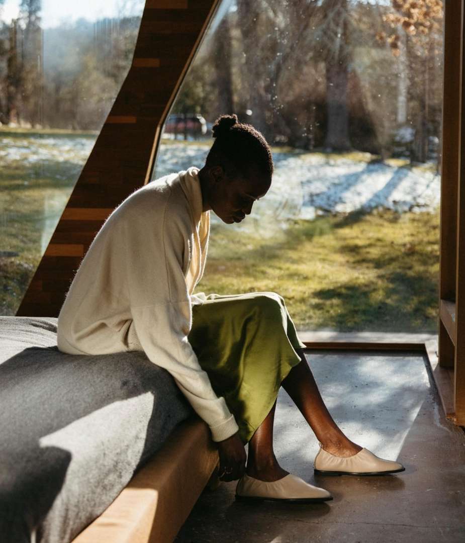 Woman in casual attire sitting on a bed in a modern house, wearing slip-on shoes, with natural light streaming through the windows.