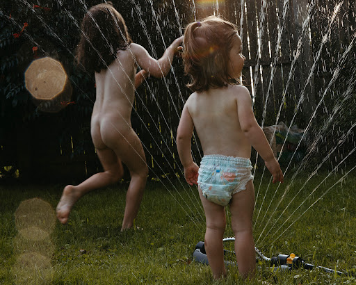 Two young children play in a garden sprinkler on green grass, bathed in bright sunlight, creating a joyful summer scene.