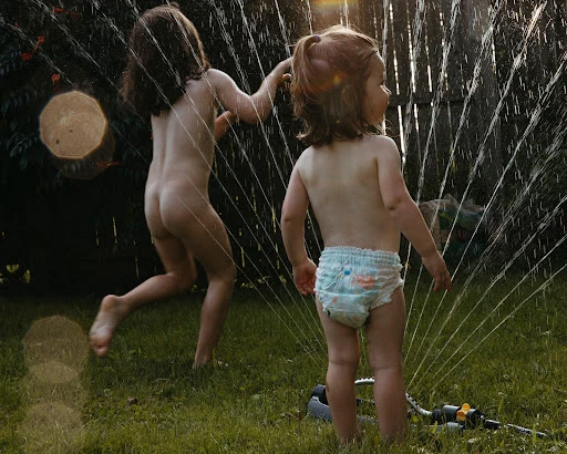 Two young children play in a garden sprinkler on green grass, bathed in bright sunlight, creating a joyful summer scene.