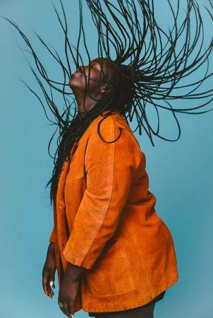 Black woman with long braided hair tossing her head back, wearing an orange suede jacket against a solid blue background.