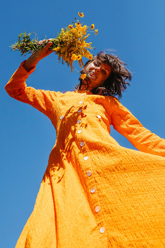 Woman in orange dress holds wildflowers against clear blue sky. Worm's-eye view capturing fashion and natural elements in bright daylight.