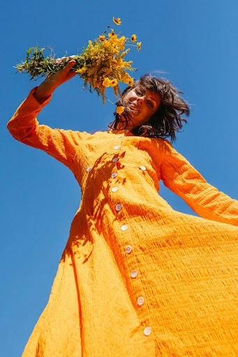 Woman in orange dress holds wildflowers against clear blue sky. Worm's-eye view capturing fashion and natural elements in bright daylight.