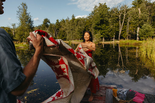 Two people hold a red and white star quilt on a wooden dock over a calm lake surrounded by lush green trees and a clear blue sky.