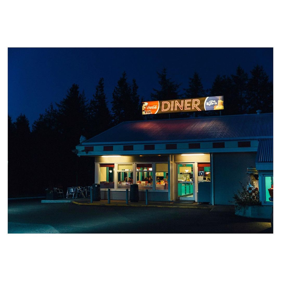 Exterior shot of a vintage diner at night with Coca-Cola sign, surrounded by dark trees. A nostalgic glimpse into roadside Americana.