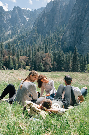 Five adults relax on a grassy field in a mountain valley. Trees dot the landscape, and rugged mountain peaks rise in the background.