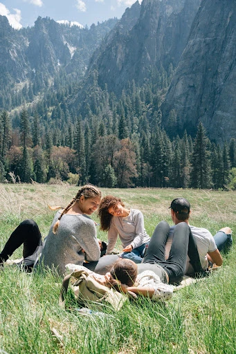 Five adults relax on a grassy field in a mountain valley. Trees dot the landscape, and rugged mountain peaks rise in the background.