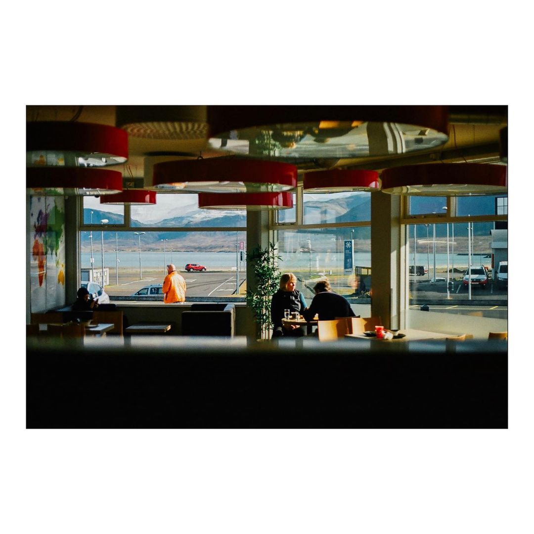 People inside a cafe look out at a scenic view of mountains, water, and sky; a car and person in orange are visible in the background.