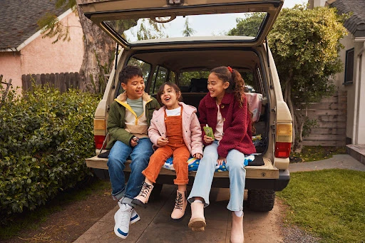 Three children sitting in the open trunk of a parked SUV, laughing and talking together.