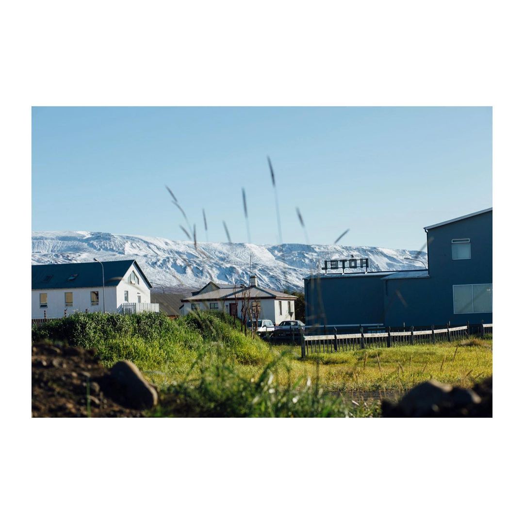 Daytime view of a hotel building and a couple of houses on a green field, against snowy mountains under a clear blue sky.