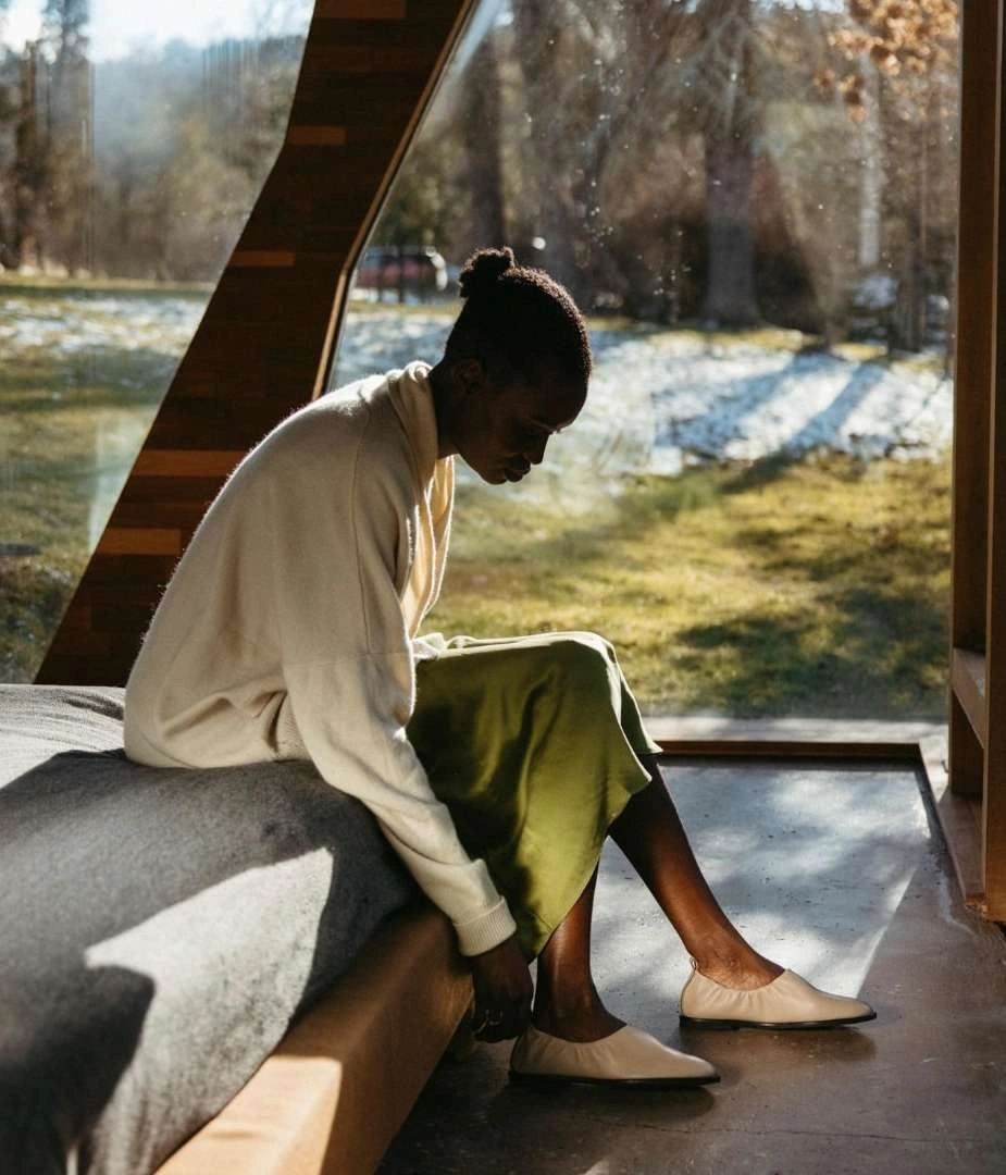 Black woman seated on a bed wearing a cream sweater and olive skirt, putting on light-colored flats
