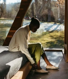 Black woman seated on a bed wearing a cream sweater and olive skirt, putting on light-colored flats