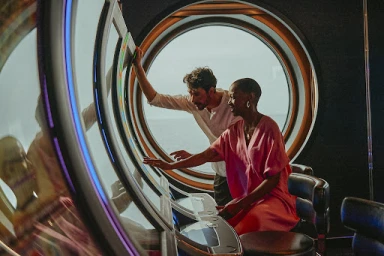 Man and woman interacting with interactive screens near large circular windows overlooking the ocean