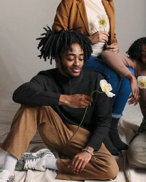 Group portrait featuring a young black man with locs holding a white flower, seated on a neutral backdrop with two women.