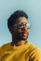 Black man wearing round sunglasses and a yellow knitted sweater against a clear sky background.