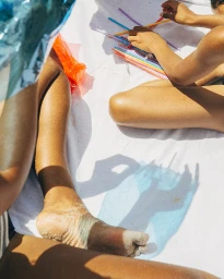 Close-up overhead shot of two people sitting on a white sheet playing with colourful plastic straws, by Emily Winiker.