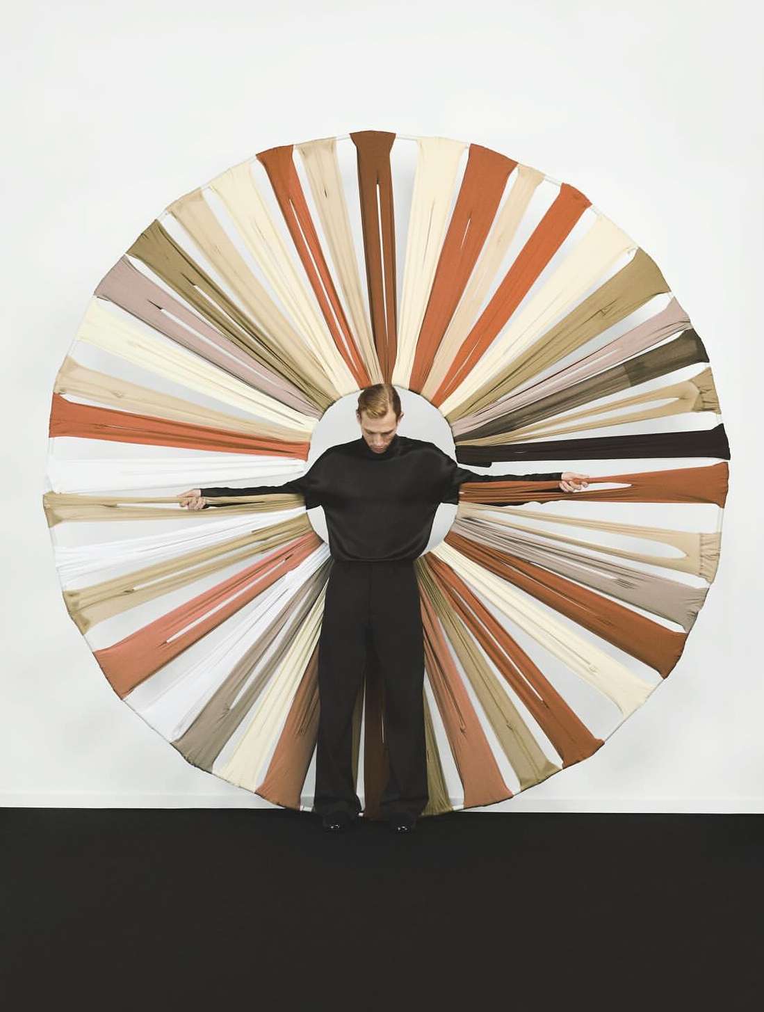Full shot of adult male posing with arms outstretched, wearing black top and pants against round structure with tan and brown fabric pieces.