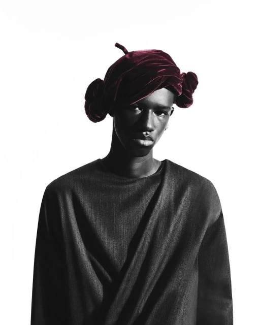 Headshot of Black model in a draped gray top and burgundy velvet head wrap against a white backdrop in a studio with dramatic lighting.