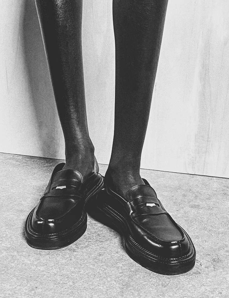 Black and white fashion shot of a Black model wearing shiny black leather penny loafers, posing in a photo studio with a neutral background.