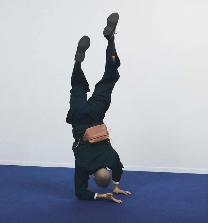 A man performing a handstand on a deep blue carpet wearing a dark suit and carrying a tan waist bag