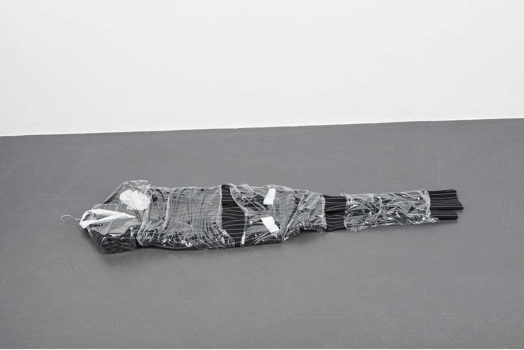 Pinstripe suit wrapped tightly in clear plastic packaging resting flat on a dark grey floor against a white wall