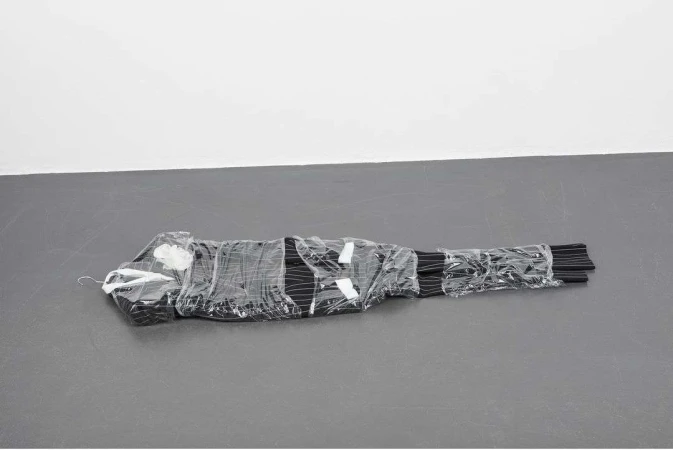 Pinstripe suit wrapped tightly in clear plastic packaging resting flat on a dark grey floor against a white wall
