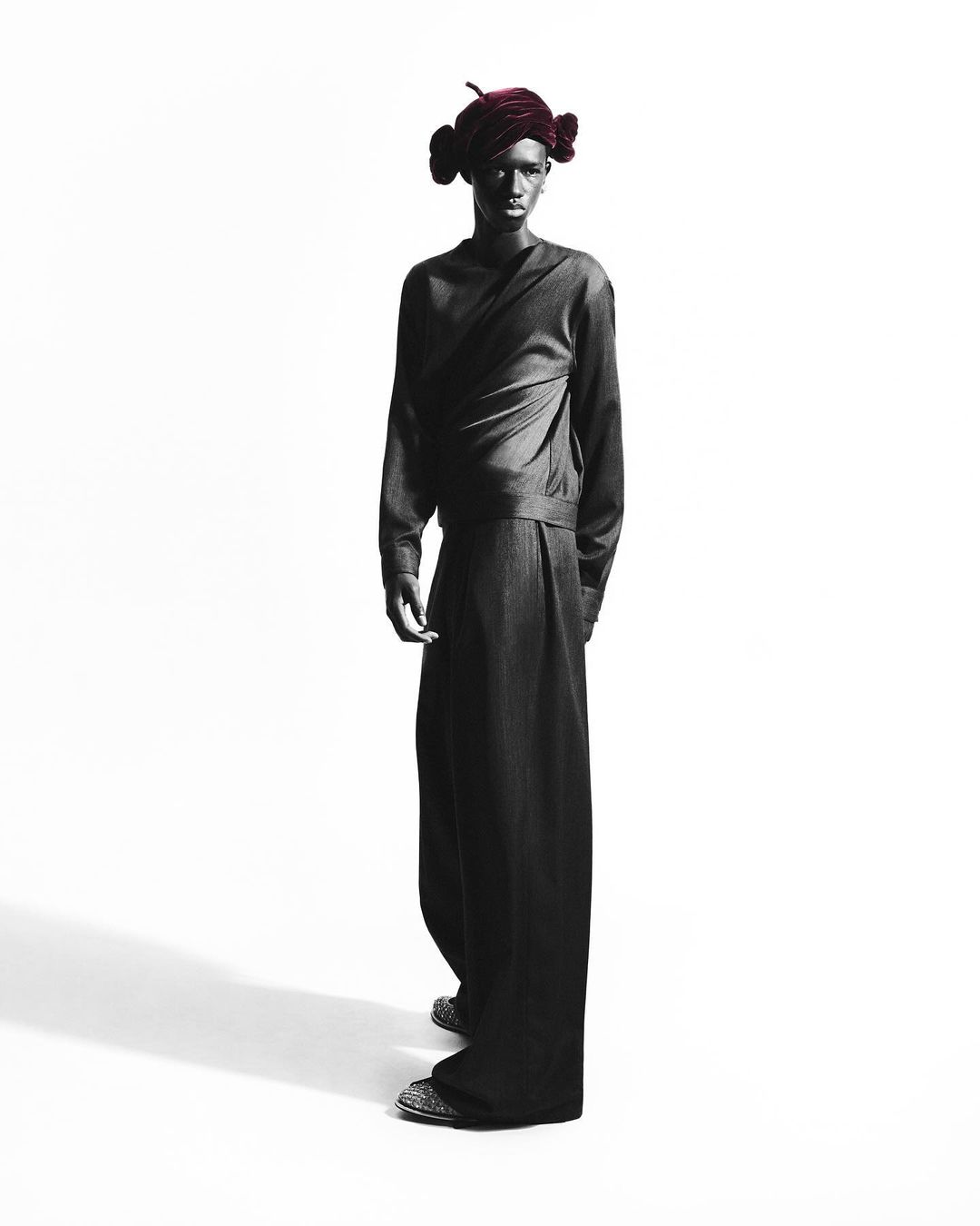 Fashion shot of a slender person with dark skin wearing a matching dark top and very wide leg pants, a dark burgundy head wrap, in a studio.
