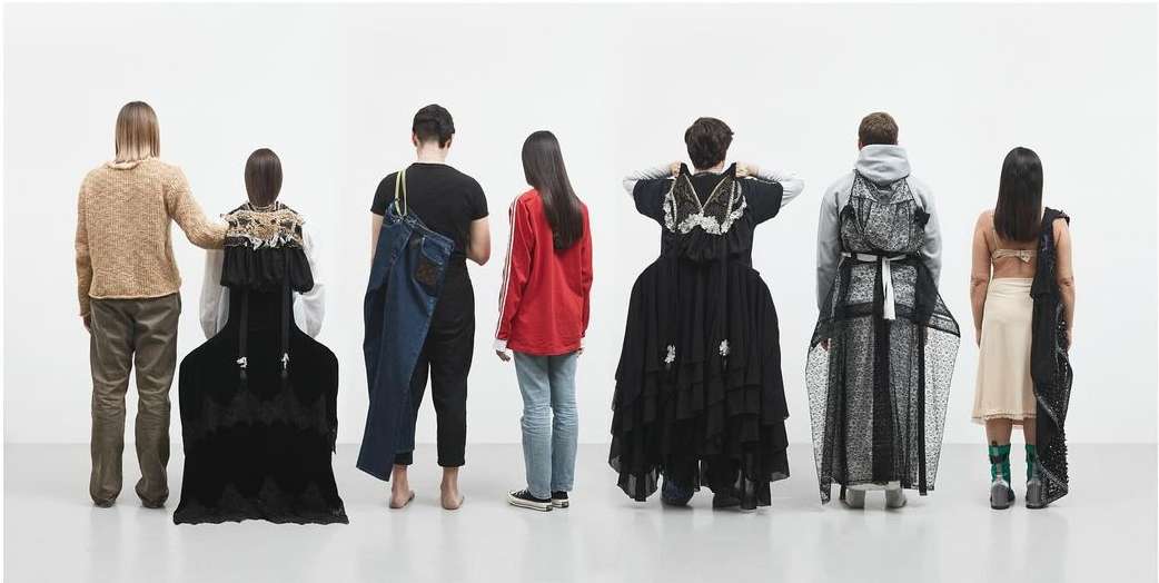 Seven people in a row facing away from the viewer in a bright white studio, wearing unique and artistic avant-garde fashion designs.