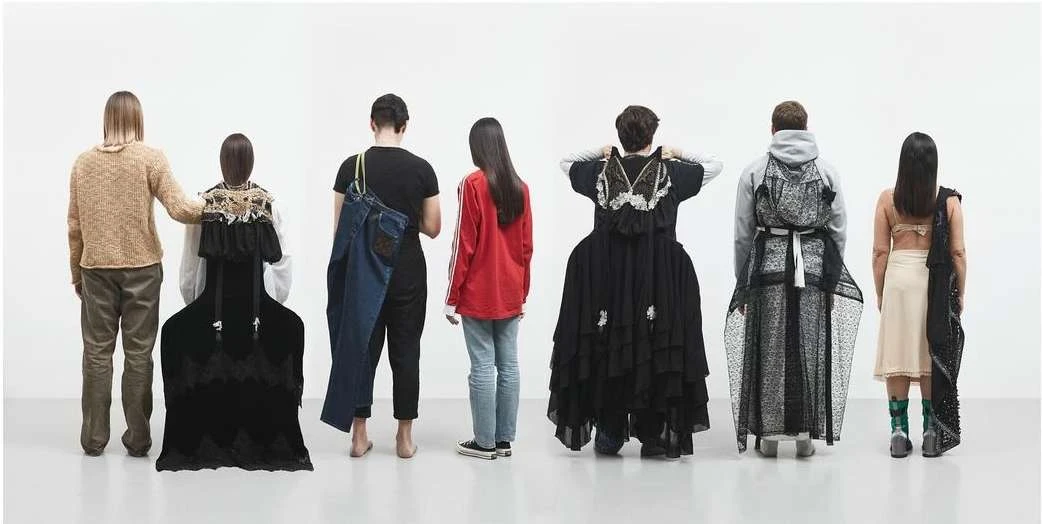 Seven people in a row facing away from the viewer in a bright white studio, wearing unique and artistic avant-garde fashion designs.