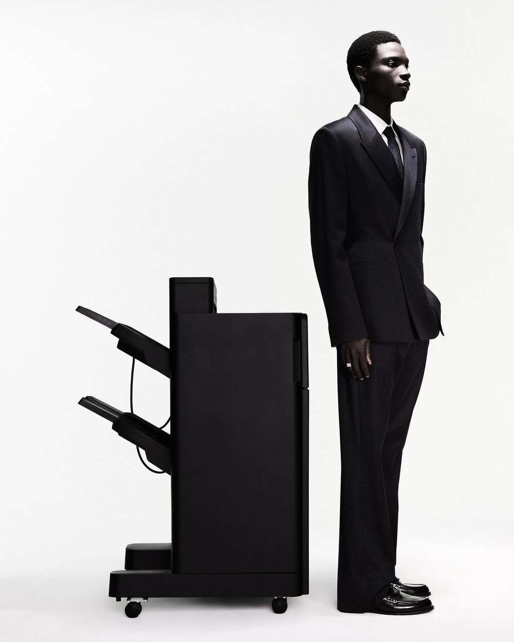 Profile shot of a dark-skinned man in a suit standing next to a black printer against a white background in a photo studio.