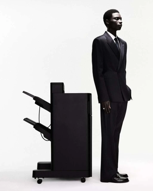 Profile shot of a dark-skinned man in a suit standing next to a black printer against a white background in a photo studio.