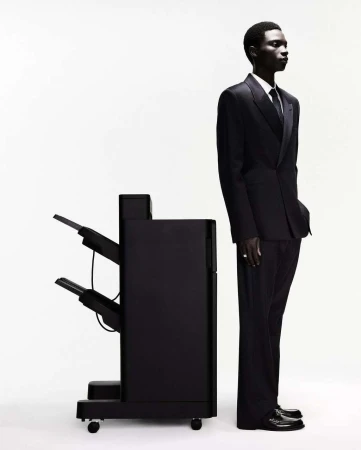 Profile shot of a dark-skinned man in a suit standing next to a black printer against a white background in a photo studio.