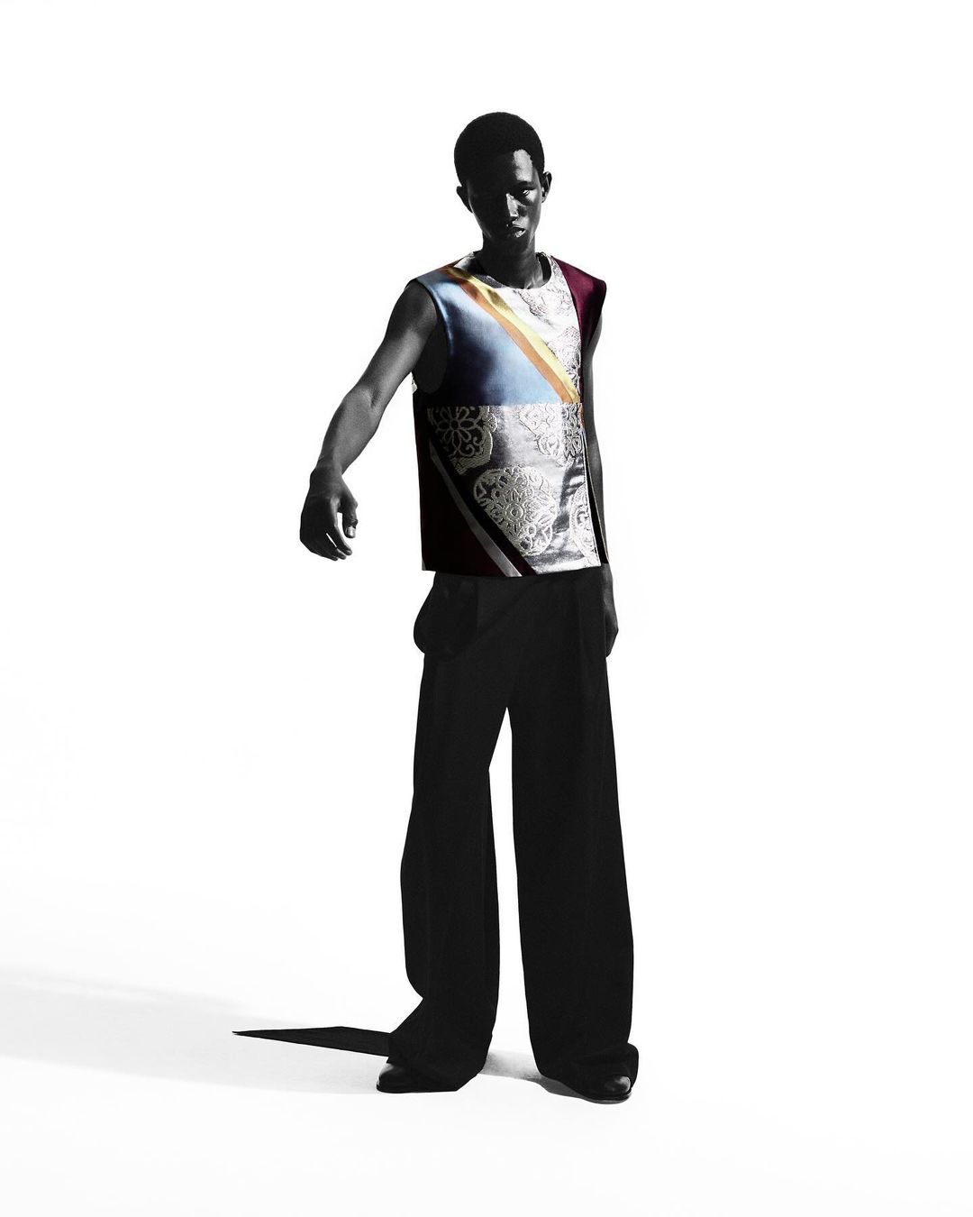 A black male model poses in a sleeveless top with wide-leg pants against a white background, casting a shadow to the lower left.
