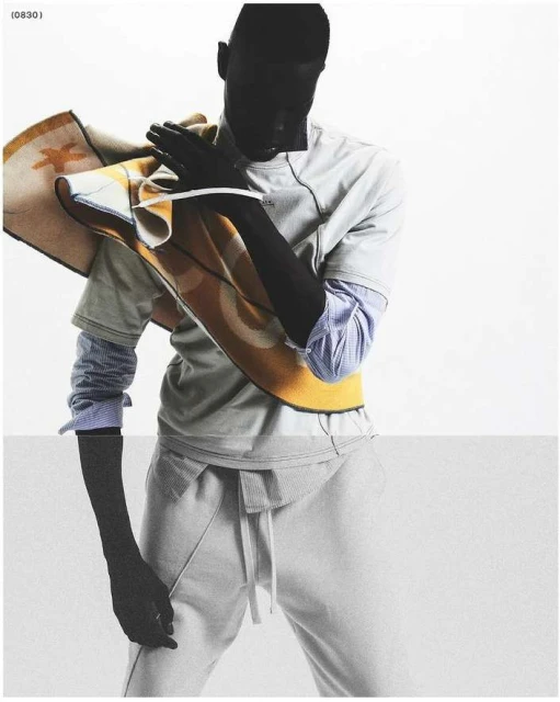 Studio shot of a male model in a gray outfit holding a patterned blanket over his shoulder against a split white and gray background.