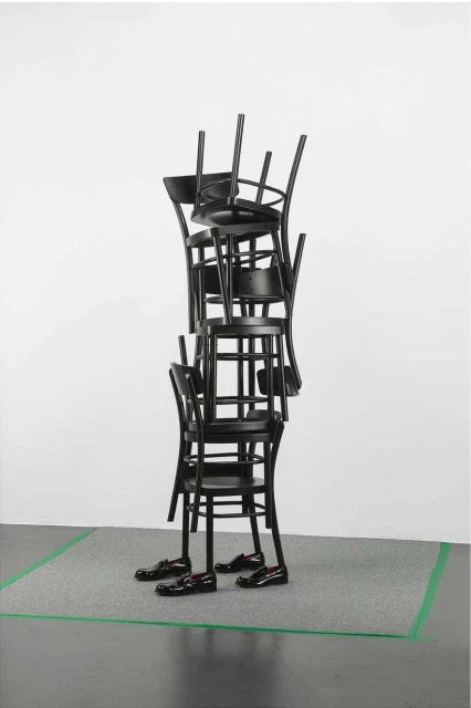 Black chairs stacked in a column wearing leather shoes with a rug under them in a studio setting with white walls and black floor.