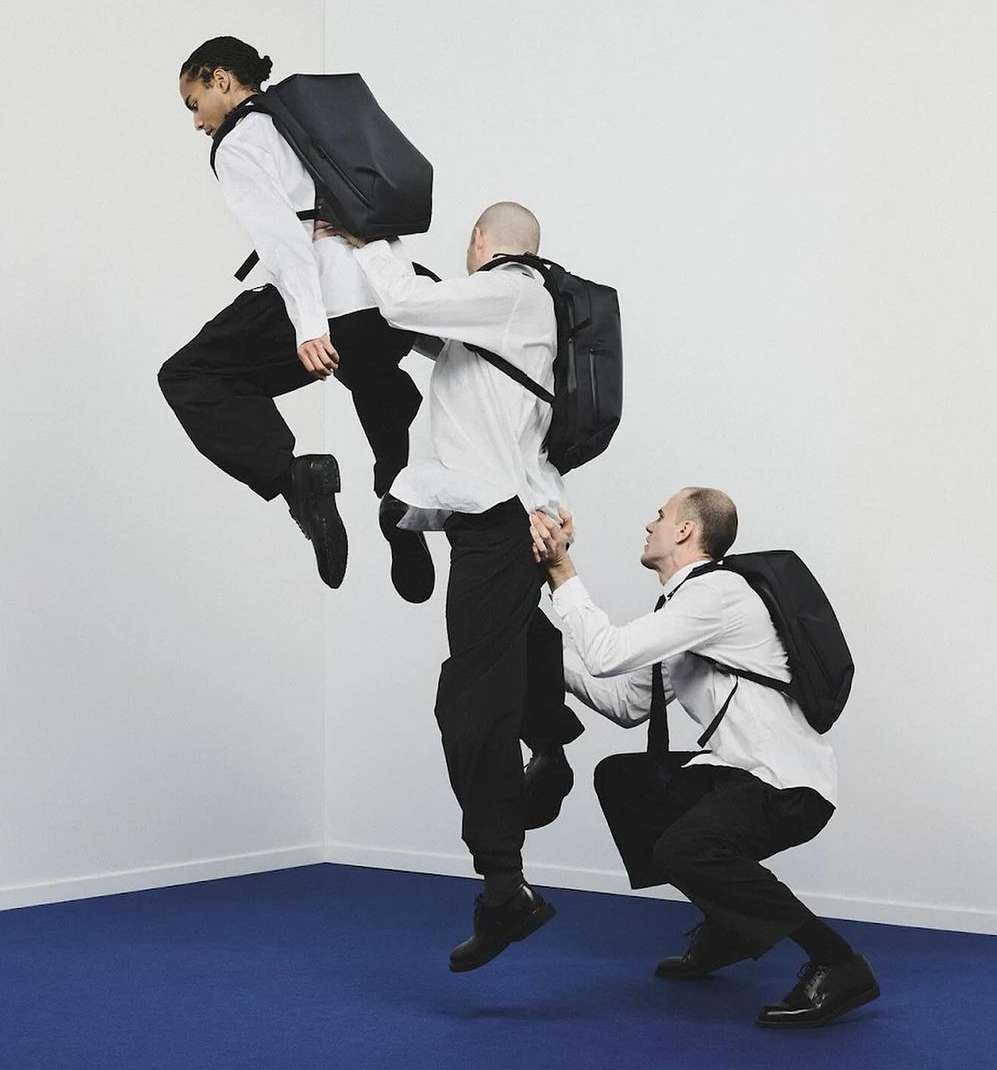 Three men in white shirts and dark pants arranged vertically; one man jumps, another supports him, set against white and blue.