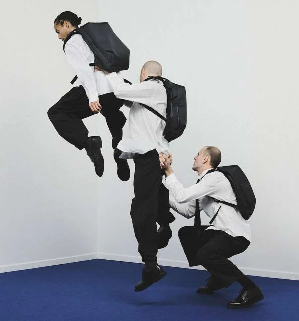 Three men in white shirts and dark pants arranged vertically; one man jumps, another supports him, set against white and blue.